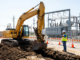 ellow excavator digging a trench near an energized electrical substation with a safety spotter directing the work.